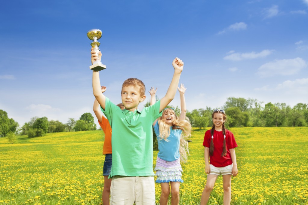 Portrait of happy little boy holding prize in lifted hands cup with his team cheering on background