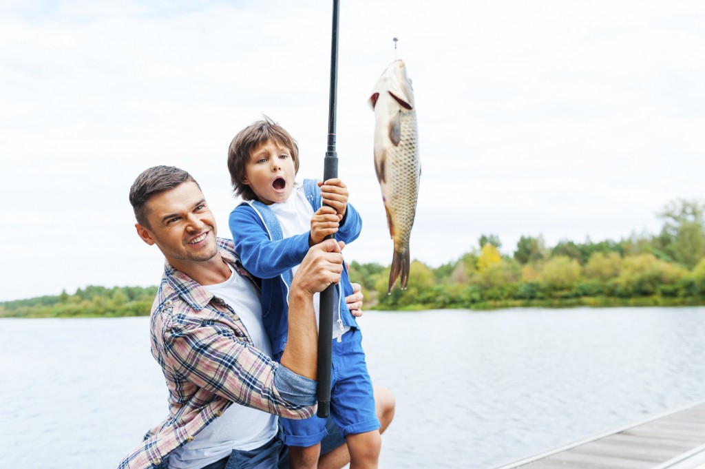 father and son stretching a fishing rod with fish on the hook while little boy looking excited and keeping mouth open
