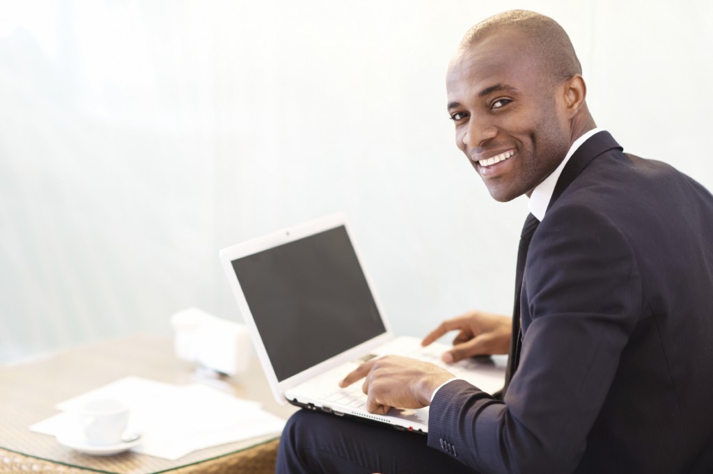 Businessman with laptop. Cheerful young African businessman typing something on laptop and looking over shoulder