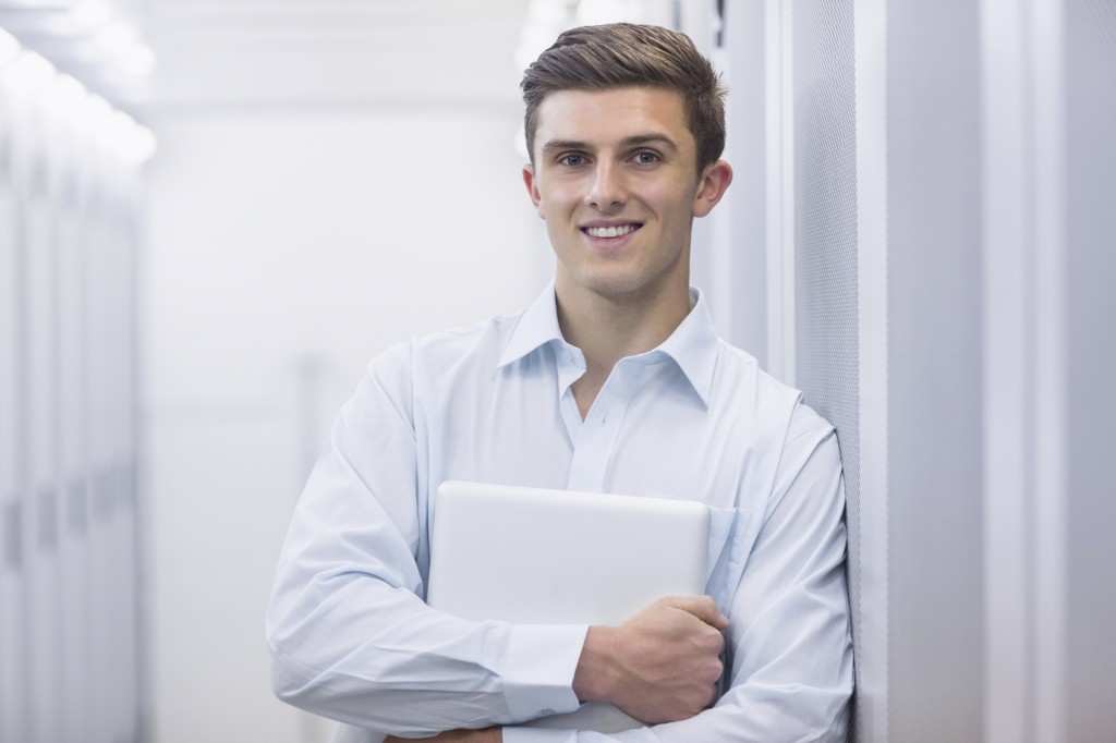 Portrait of a smiling technician holding a laptop and leaning against a tower in a large data center