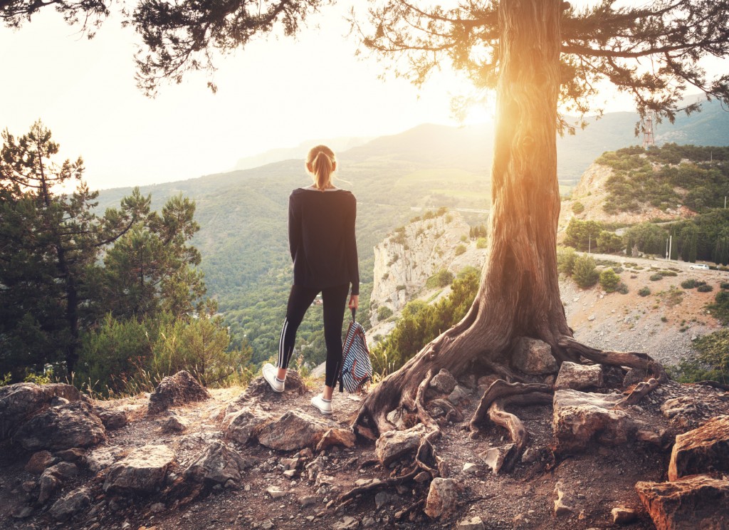 Young woman standing on the mountain near old tree at sunset.