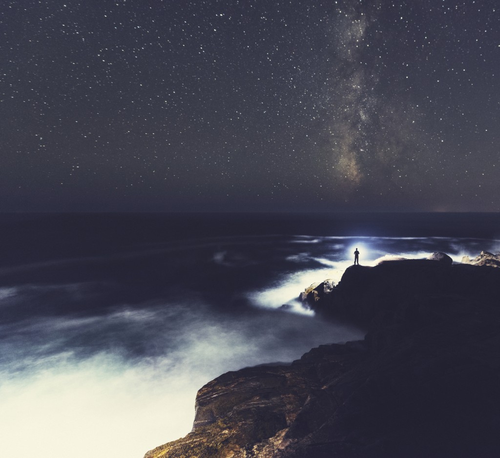 A man stands at the edge of a cliff shining a bright light out into the Atlantic while the Milky Way arcs high above.