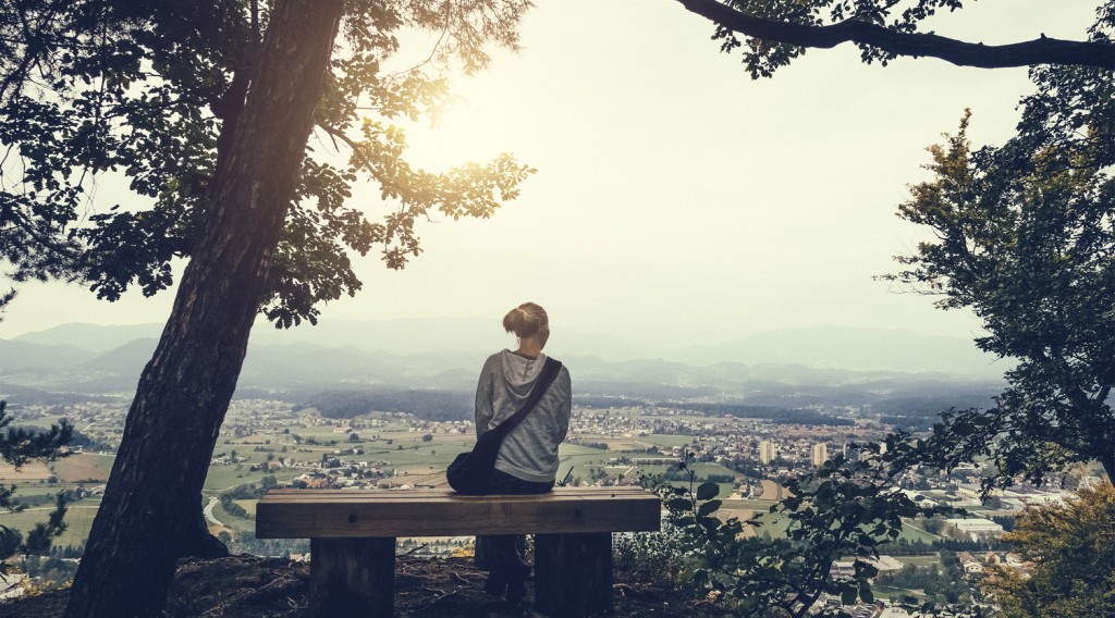 Young woman sitting on bench on top of the hill looking down on her home town.
