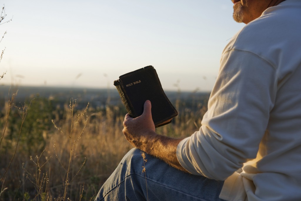 Man praying while holding a Holy Bible in an open field.