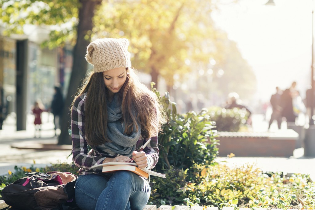 Student girl writing in notebook and studying. Casual clothing, scarf and hat.