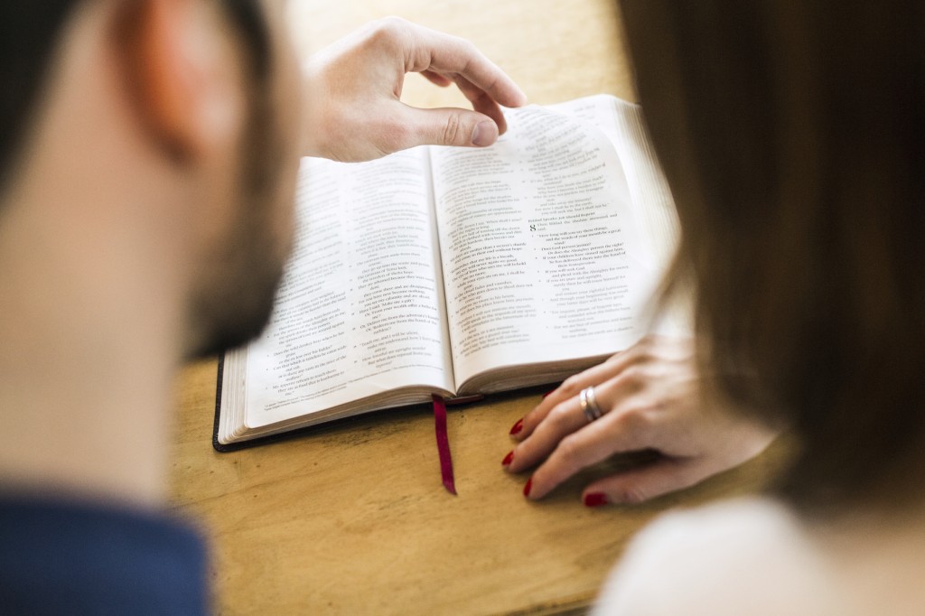 An adult man and woman study the Bible together, sharing their favorite scriptures while enjoying a cup of coffee. The husband reads aloud while pointing out a verse to his wife. Shot as viewed from behind and over their shoulders, the focus on the scripture in the background.