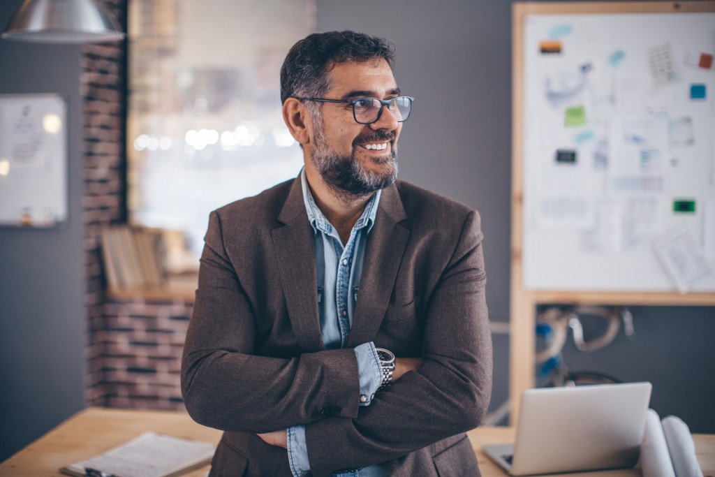 Mature business man working in modern office. Standing in front office and looking away witn his arms crossed. Wearing casual clothes and glasses.