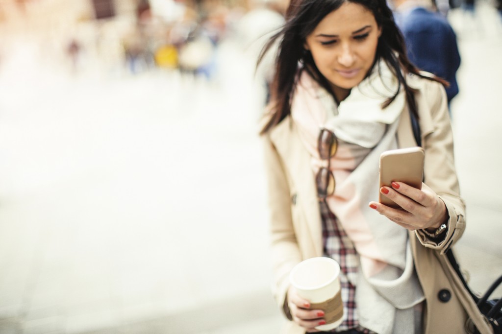 Photo of young modern woman using a smartphone outdoors on a beautiful day and holding cup of coffee in other hand