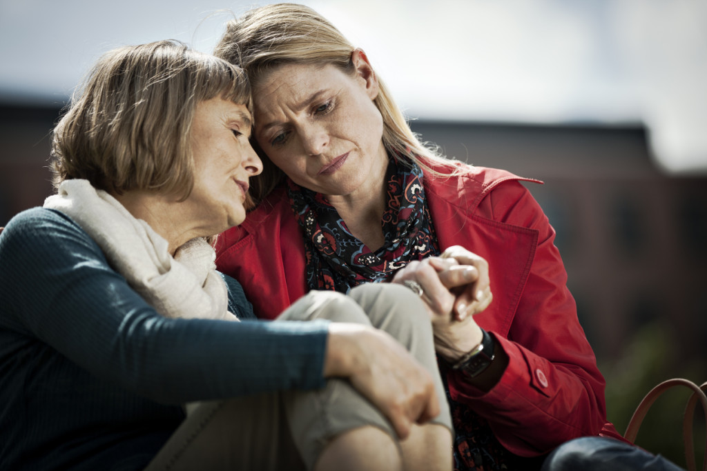Mature woman and her adult daughter sitting close to each other with sad expression on their faces. Younger woman is holding her mother's hand.