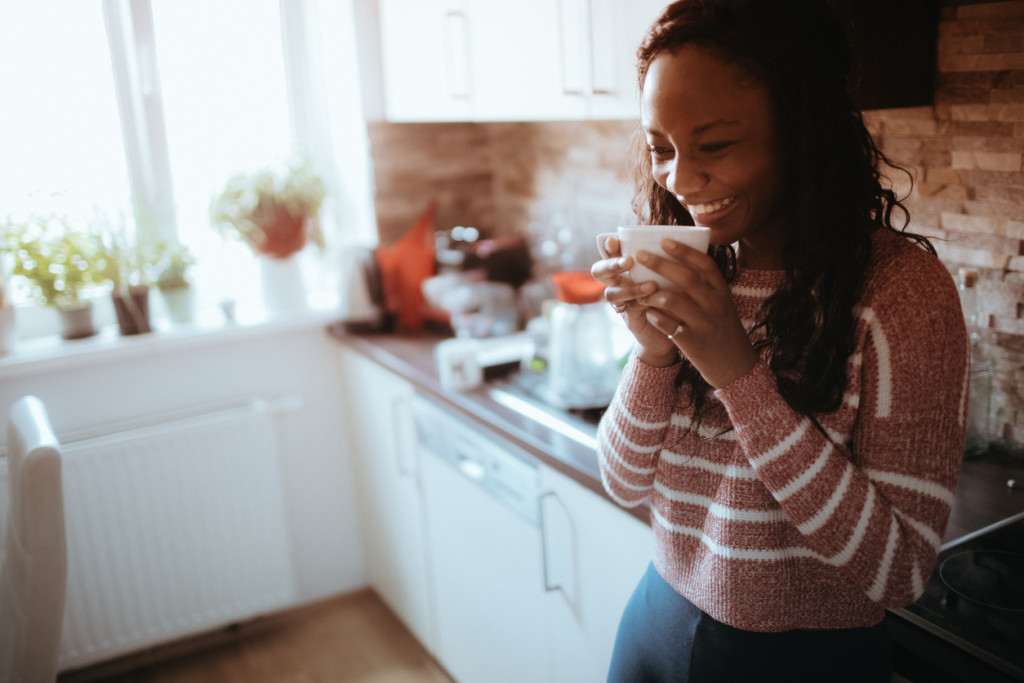 Smiling young woman enjoys cup of coffee in her kitchen, early in the morning