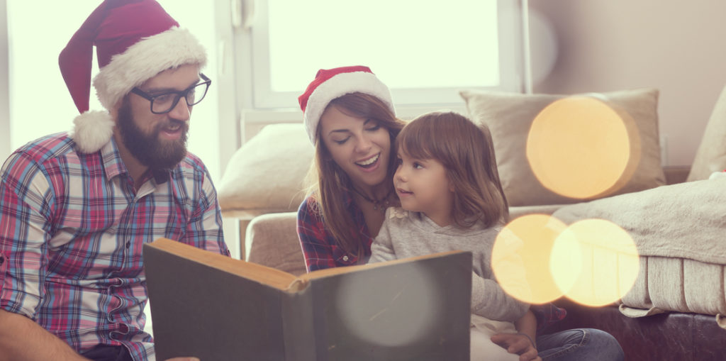 Young parents sitting on the living room floor next to a nicely decorated Christmas tree, mother holding a baby girl in her lap and all together reading a fairy tale. Focus on the baby girl