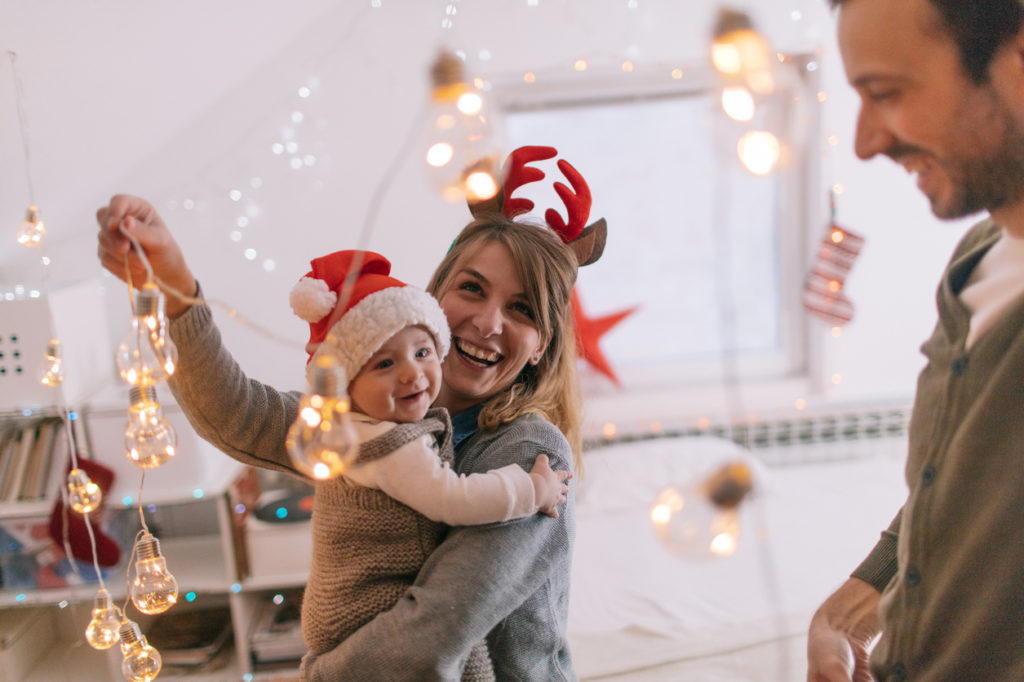 Photo of a cheerful young family with one child, decorating for the first Christmas they are spending together with a baby