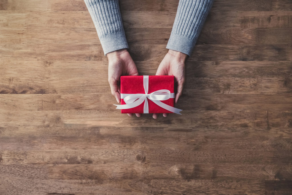 Woman hands in gray sweater on wood table giving red Christmas gift box