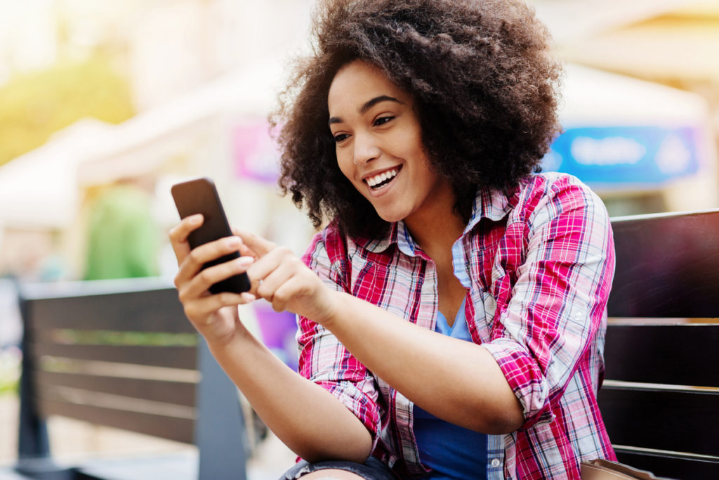 Smiling young African ethnicity woman sitting on a bench outdoors and texting.
