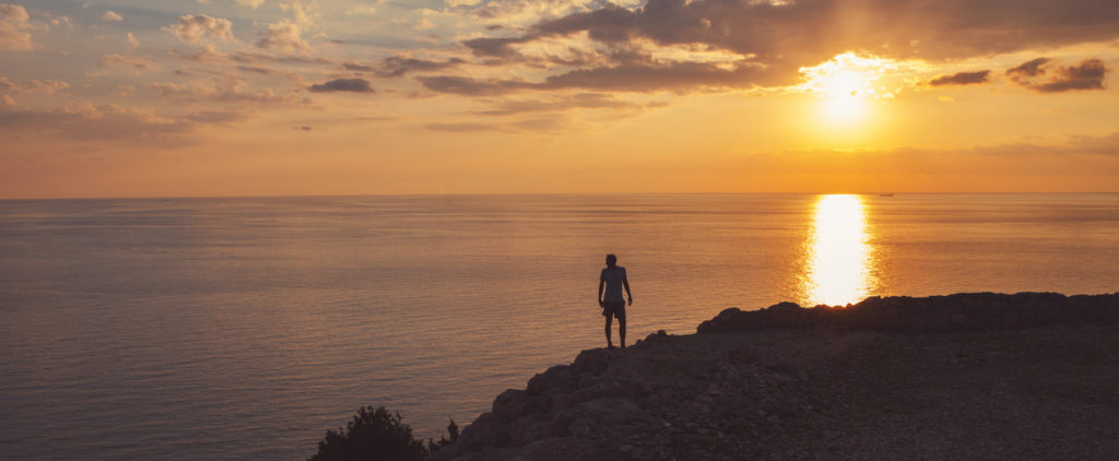 Silhouette of a man standing on the cliff and watching sunset over the sea horizon.