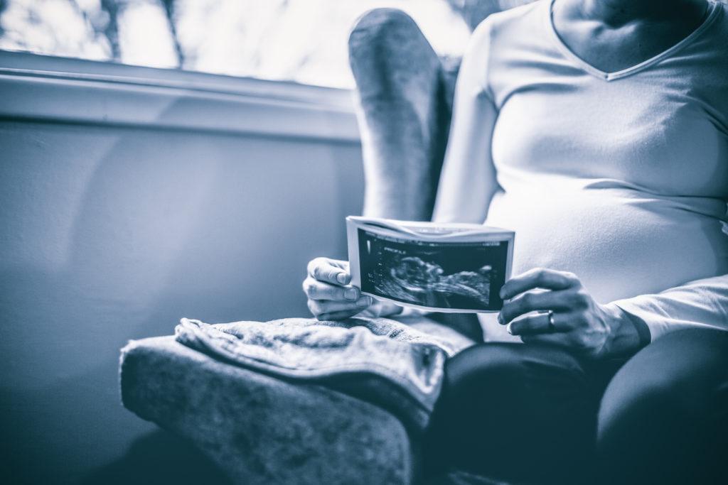 A young pregnant woman studies a picture of her in-utero baby as pictured in an ultrasound image. Horizontal with copy space. Blue toned image.