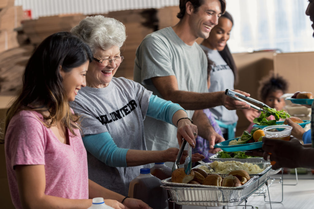 Cheerful senior woman and her friends or family serve meals to homeless people in a soup kitchen.
