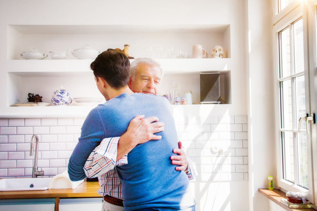 Senior man embraces mature son in sunny kitchen with a proud smile.