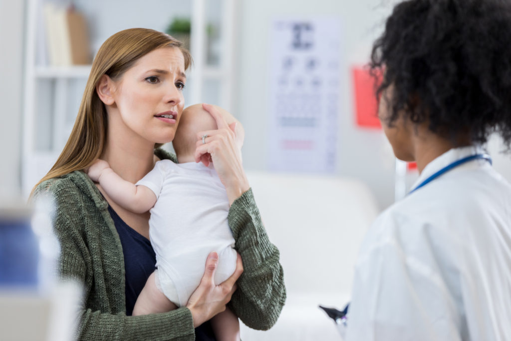 Worried new mom comforts her newborn baby while talking with the child's pediatrician.