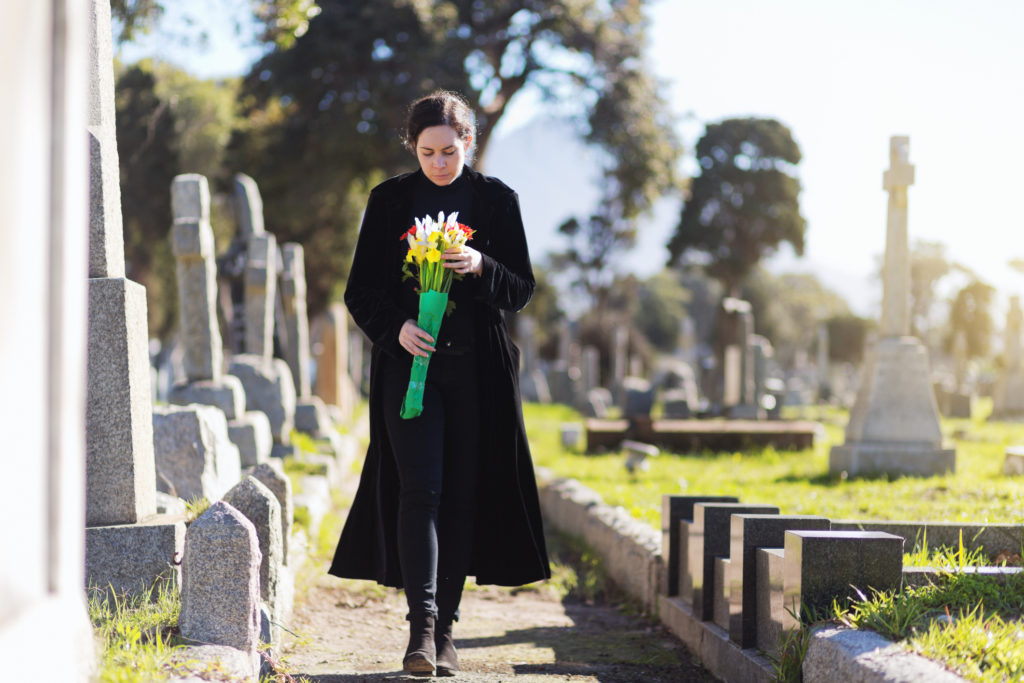 A young woman in black walks through cemetery headstones carrying flowers to the grave of someone she misses.