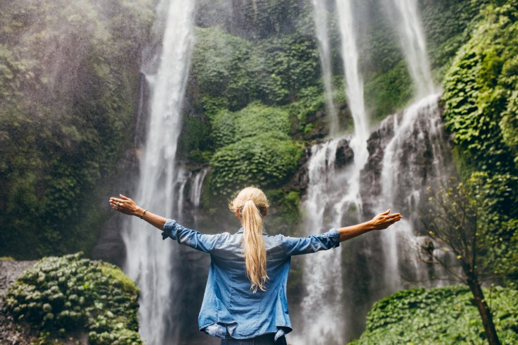 Rear view of young woman standing in front of waterfall with her hands raised.