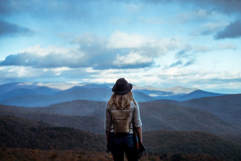 A photograph of a young woman hiking through the mountains in the fall or early winter enjoying the view.