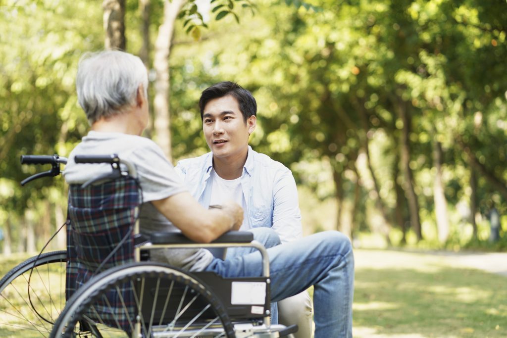 young asian adult son chatting with wheel chair bound father outdoors in park
