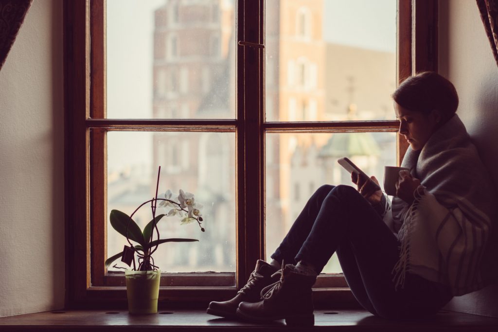 Woman on the window drinking coffee and surfing the net on smartphone