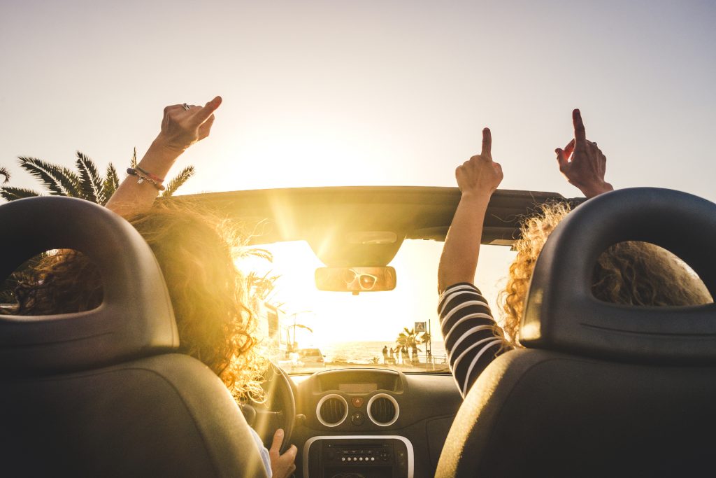 Couple of woman friends traveling and driving having a lot of fun dancing in the car with opened roof and summer vacation sunset ocean in front - concept of friendship together and nice lifestyle for independent girls