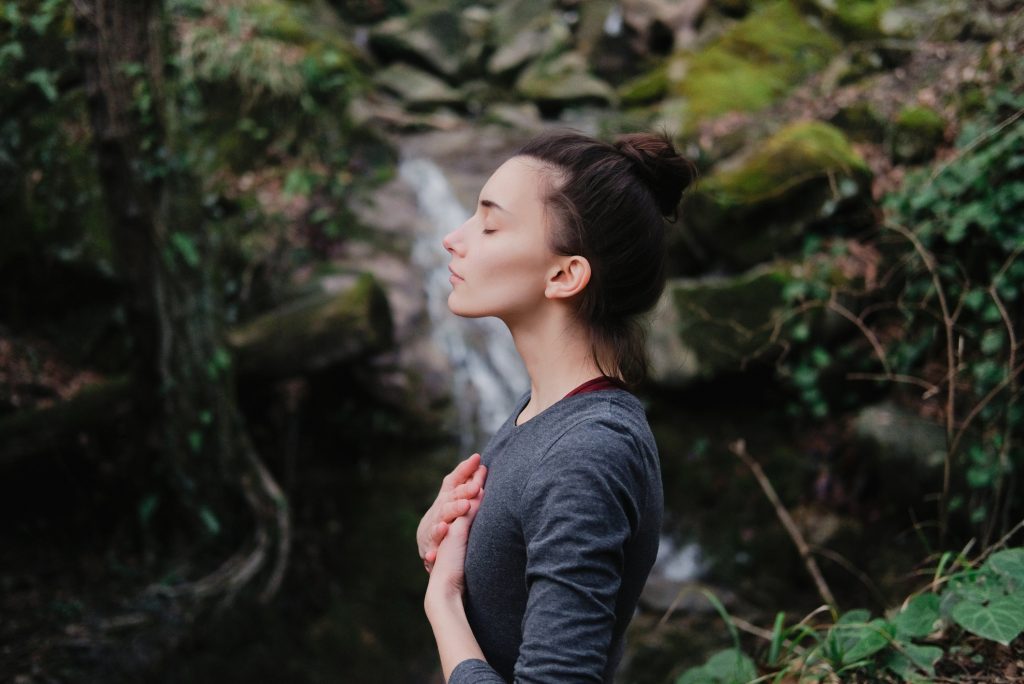 Young woman breathing deeply outdoors in moss forest on background of waterfall.