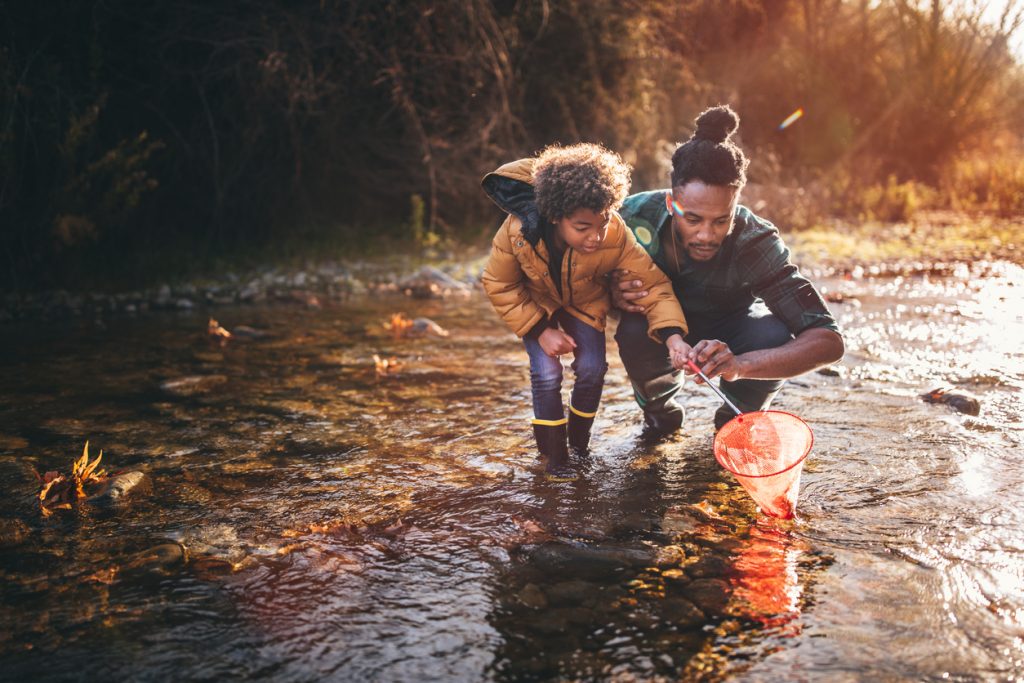 Young father teaching son how to fish with fishing net in mountain stream at sunset