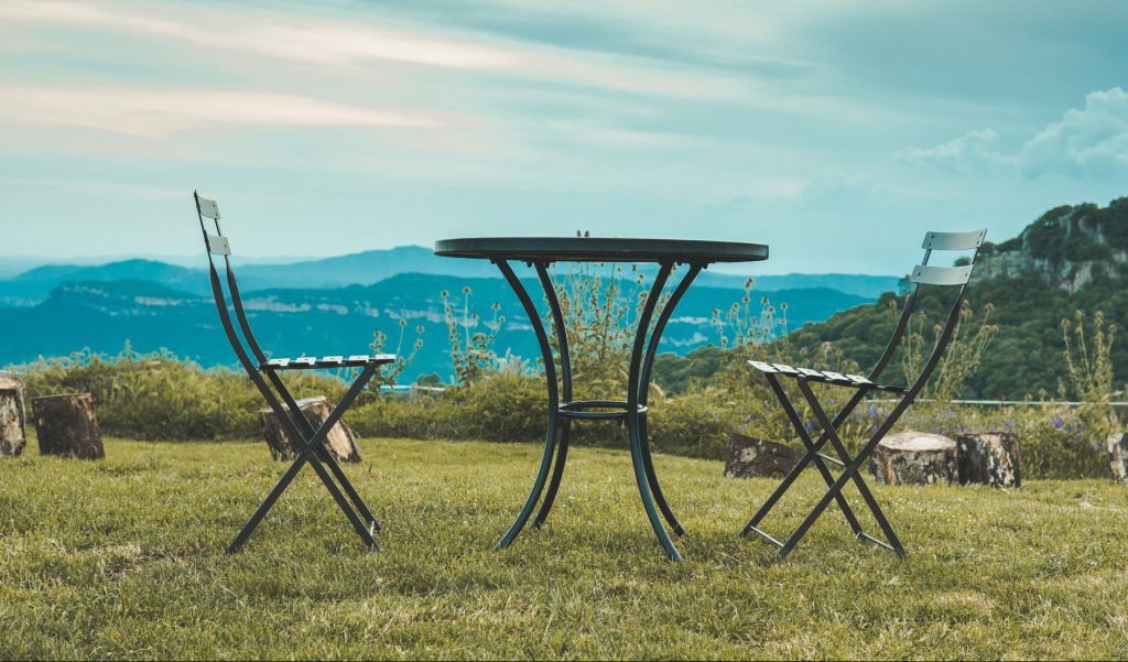 Two chairs facing each other and a table in the middle of nature. Behind it you can see the mountains and the clouds. They are high in a valley.