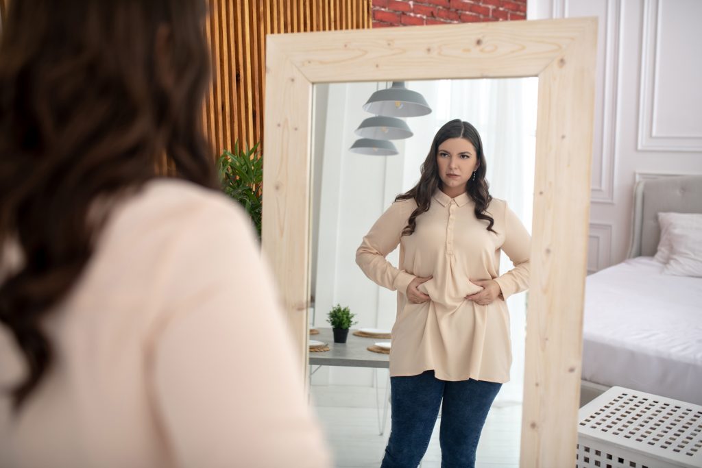 Young woman in a beige blouse squeezing fat on her belly while looking in a mirror.