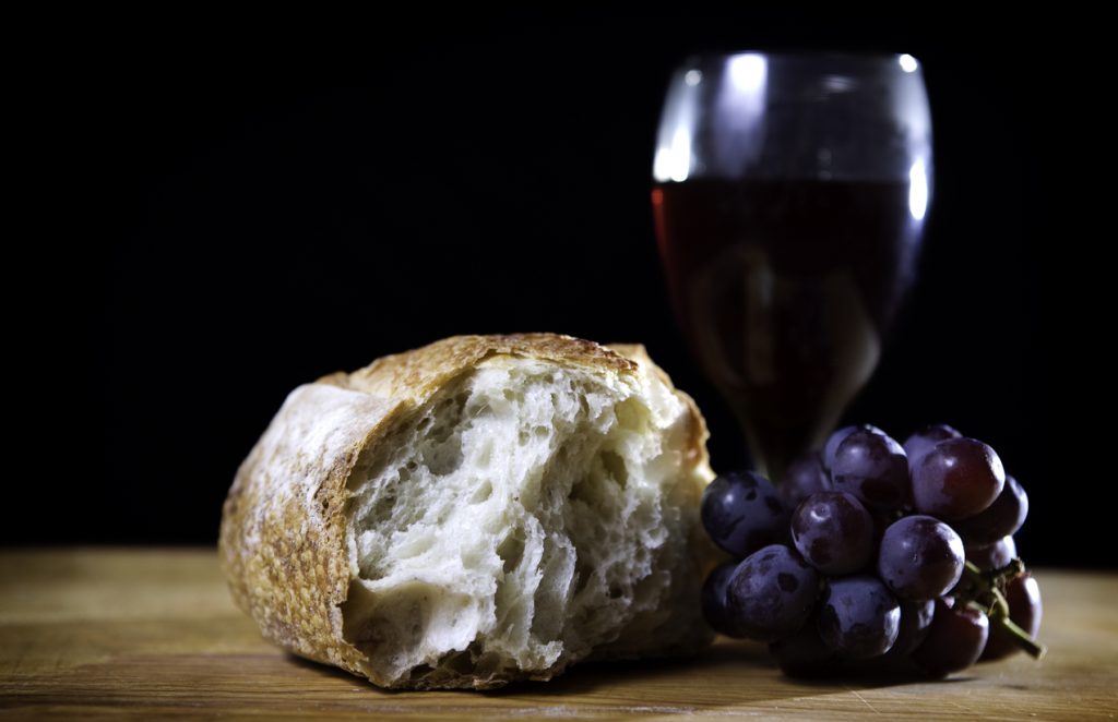 A dark high contrast portrait of bread, wine, and grapes, representing the Last Supper. Black background; horizontal with copy space.