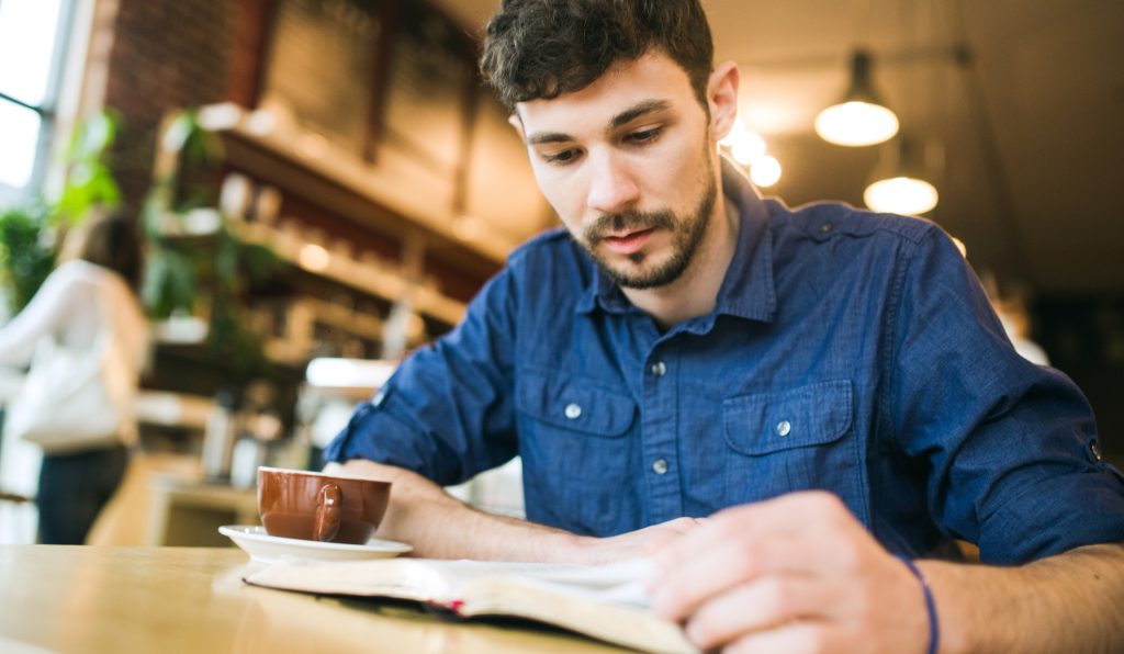 A man becomes more spiritually resilient as he reads the Bible or a book in his favorite coffee shop, a latte he's drinking sitting next too him on the table. Horizontal image with copy space.