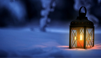 A candle-lit lantern in snow. It's night and the candle glows orange in the dark blue snowy forest.