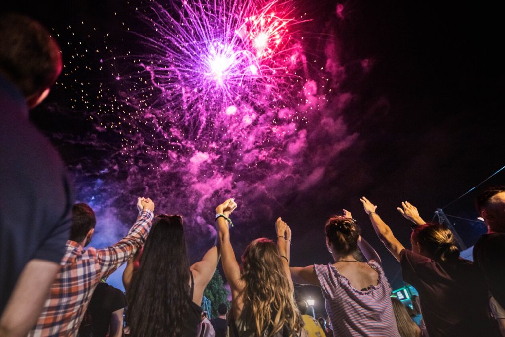 A group of friends holding hands and lifting them up as fireworks burst in the sky above them