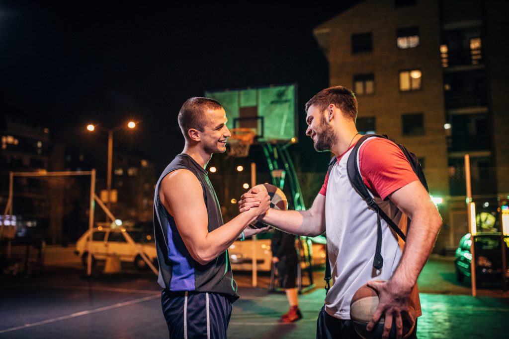 Group of men, streetball players standing on court at night downtown in city, handshake after the game.