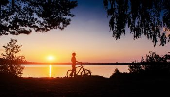 A woman walking her bike next to a lake during a sunrise